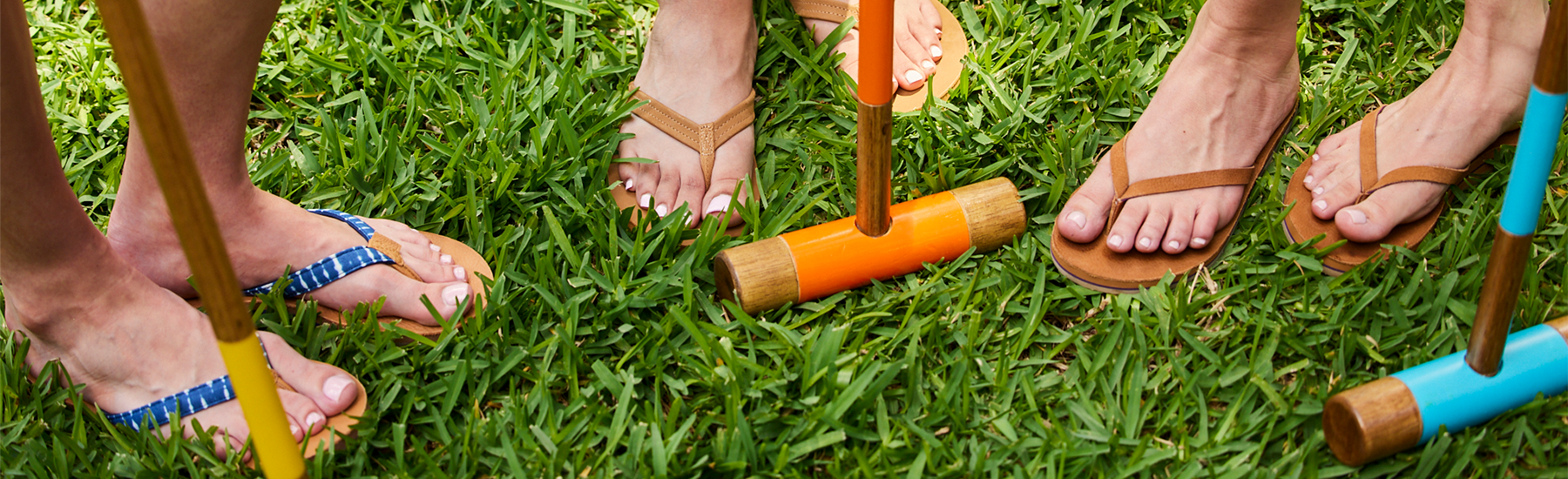 This high-angle, close-up shot focuses on the feet of four people gathered on a lush green lawn for a game of croquet. The image emphasizes a relaxed, summer social atmosphere through a mix of colorful sports equipment and casual footwear.