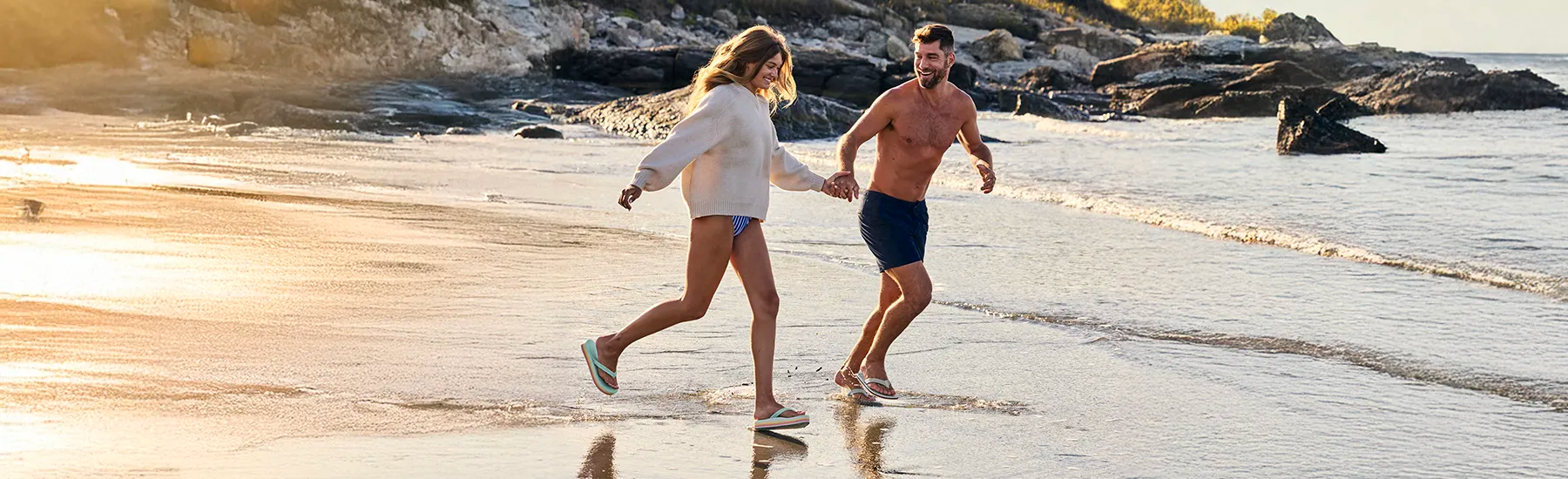 The warm, golden sunlight reflects off the water and sand, highlighting the couple's coordinated beach attire and durable summer footwear.