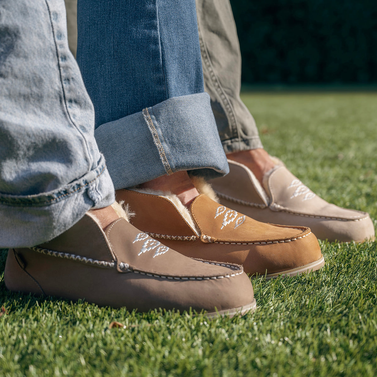 Taupe and tan Alta slippers with white embroidered details, worn with cuffed jeans on grass.