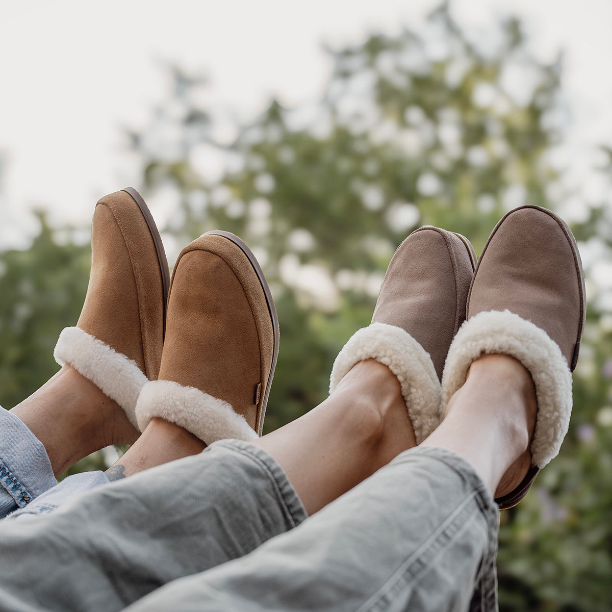 Two people wearing tan and taupe slippers with cream plush trim, resting their feet outdoors.