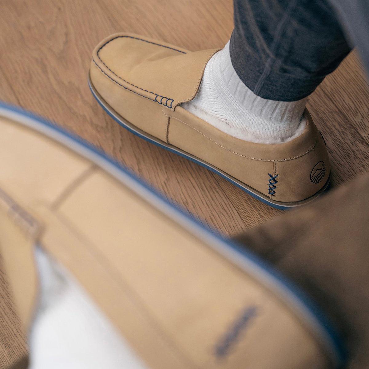 Light tan Sonoro loafer with blue contrast stitching and sole, worn with white socks on a wooden floor.