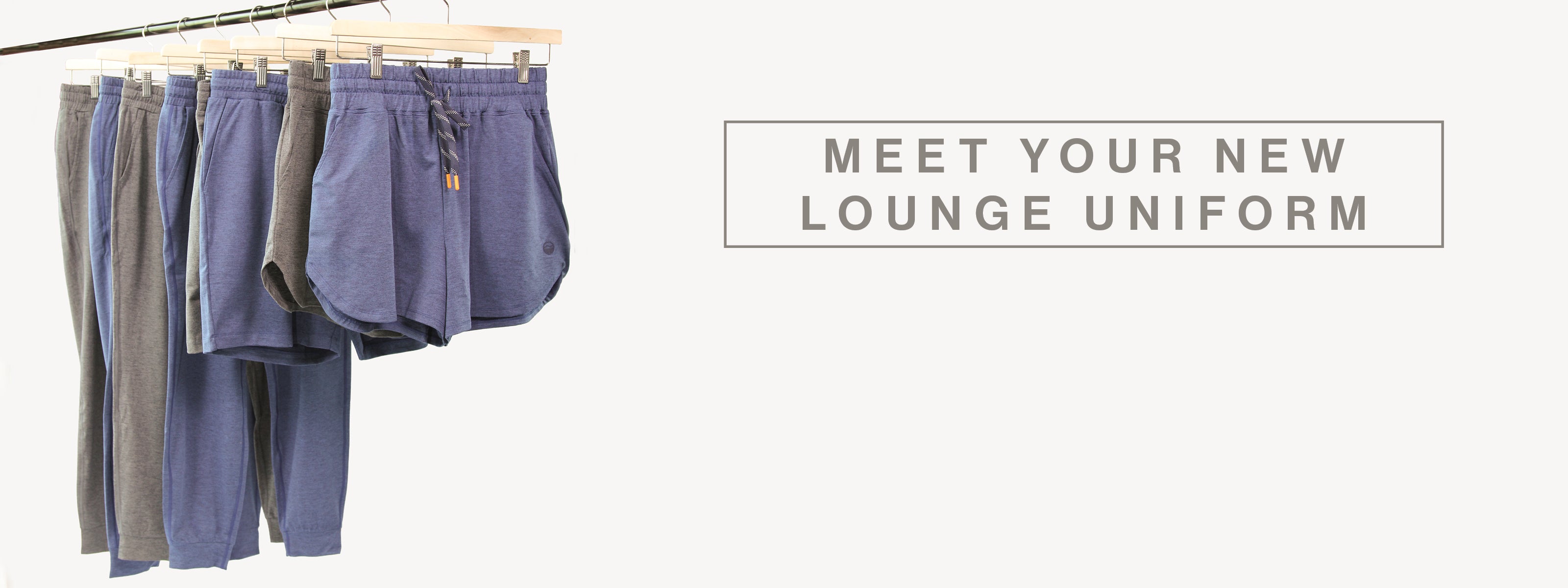A set of lounge pants in muted blue, gray, and taupe hanging on a wooden rack against a light gray background, with the text 'MEET YOUR NEW LOUNGE UNIFORM' on the right.