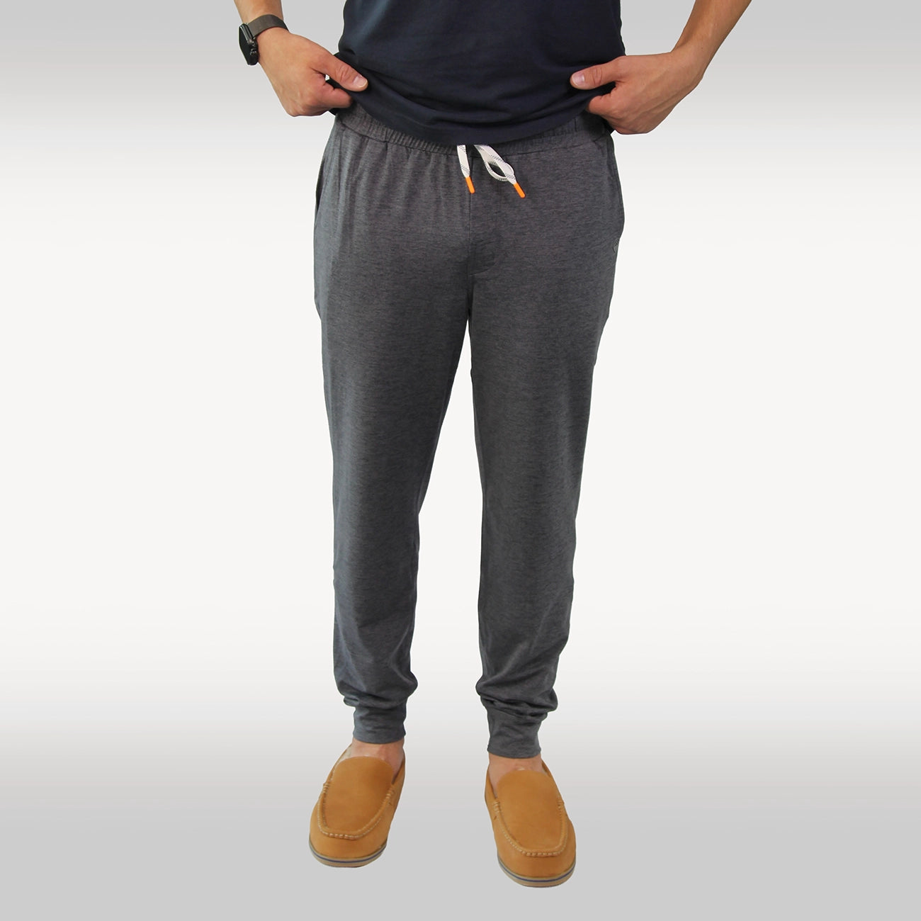A person wearing dark gray heather jogger joggers with a white drawstring, paired with a navy shirt and tan Hari Mari slippers, shown from the waist down against a light gray studio background.