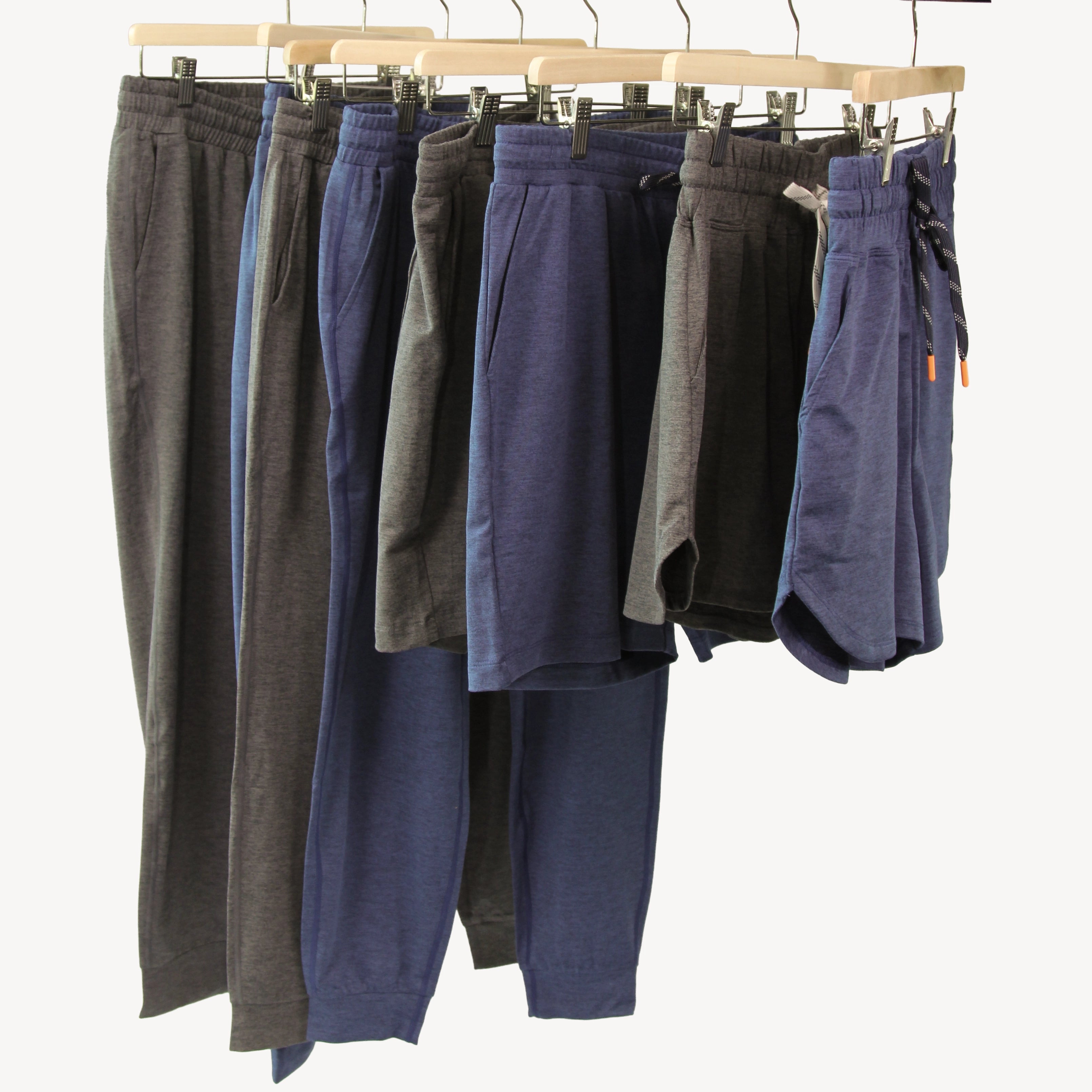 Four pairs of casual lounge bottoms from Hari Mari hanging on wooden hangers against a plain light background: two jogger pants in dark gray and navy, and two shorts in dark gray and navy, each with elastic waistbands and drawstrings.