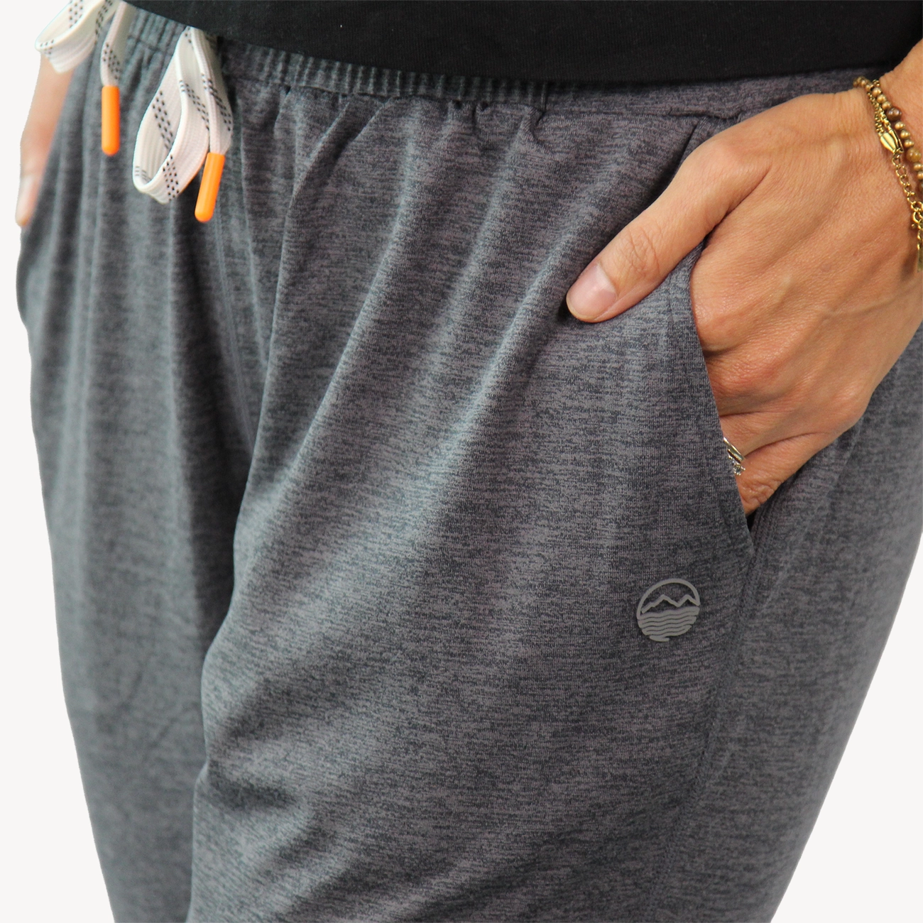Close-up of a woman wearing dark heather gray athletic joggers from Hari Mari with a side pocket, hand in pocket, elastic waistband, and white drawstrings with orange tips.