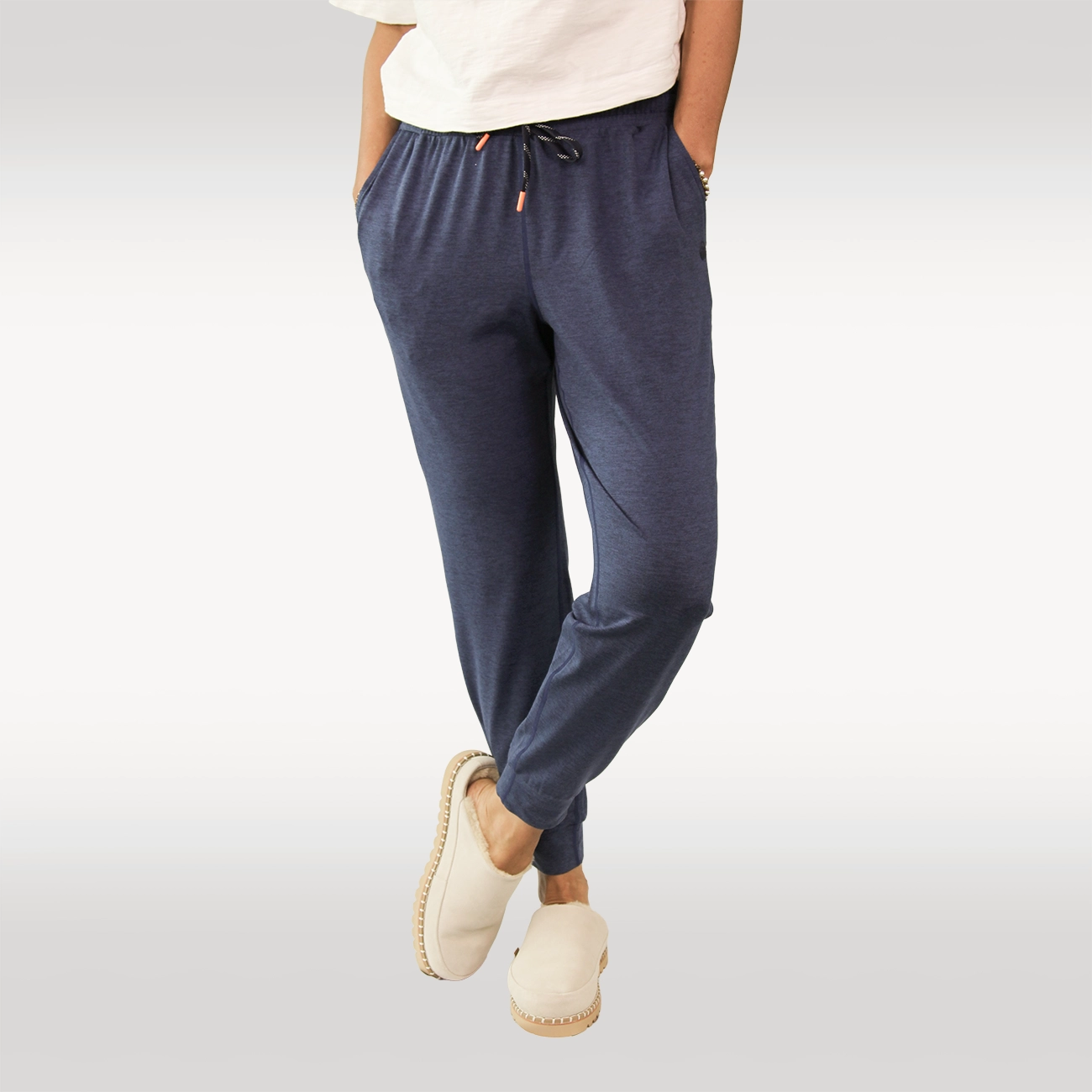 A woman wearing dark heather gray drawstring joggers with side pockets, paired with tan Hari Mari slippers and a black top, shown from the waist down against a light gray studio background.