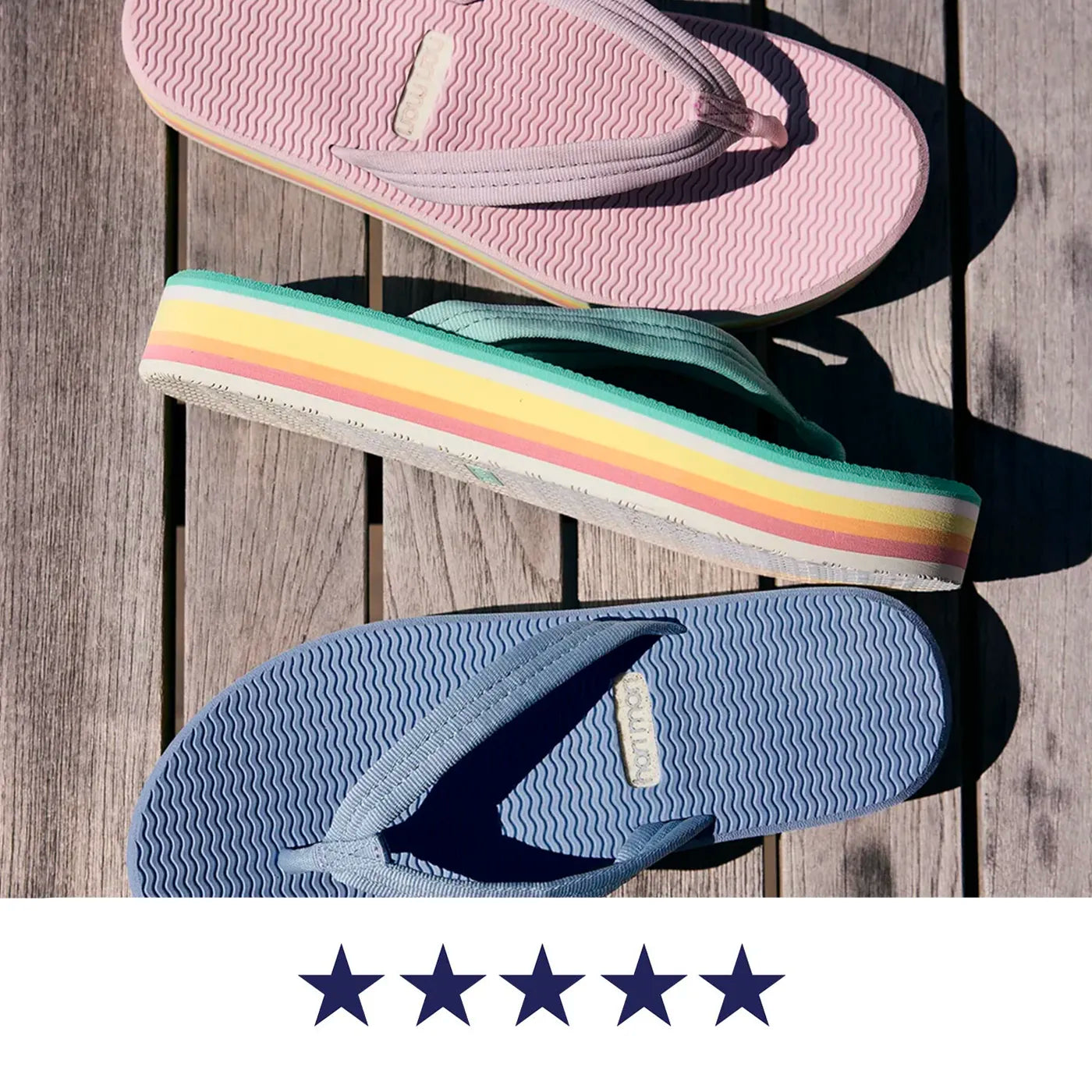Three pastel flip-flops arranged diagonally on a weathered wooden plank surface. The top flip-flop is light pink, the middle has rainbow-colored stripes along the side, and the bottom is light blue. All three are Hari Mari Dunes Sunset flip flops for women