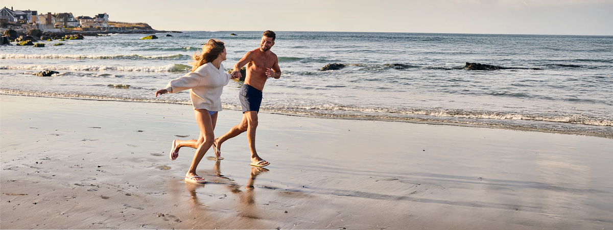 Two people running on a beach wearing Hari Mari flip flops with waves in the background