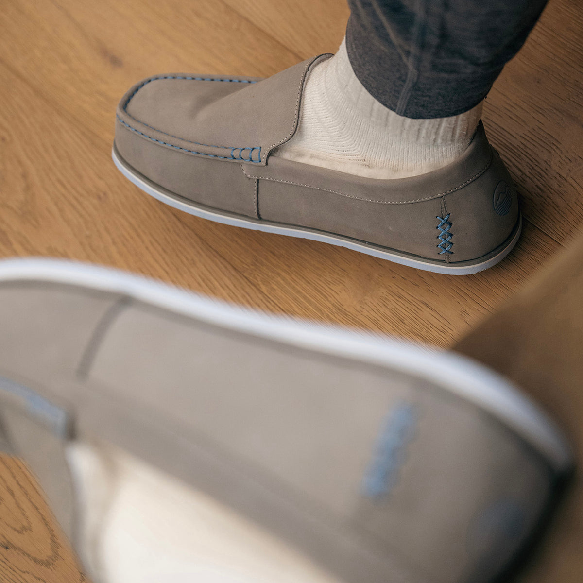 Light gray Sonoro loafer with blue contrast stitching, worn with white socks on a wooden floor.