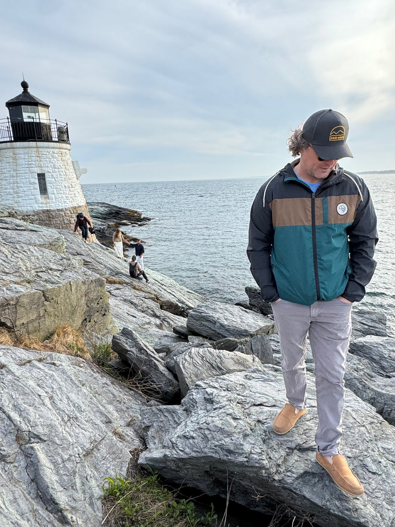 man standing on rocks next to a lighthouse and beach with photoshoot taking place in the background