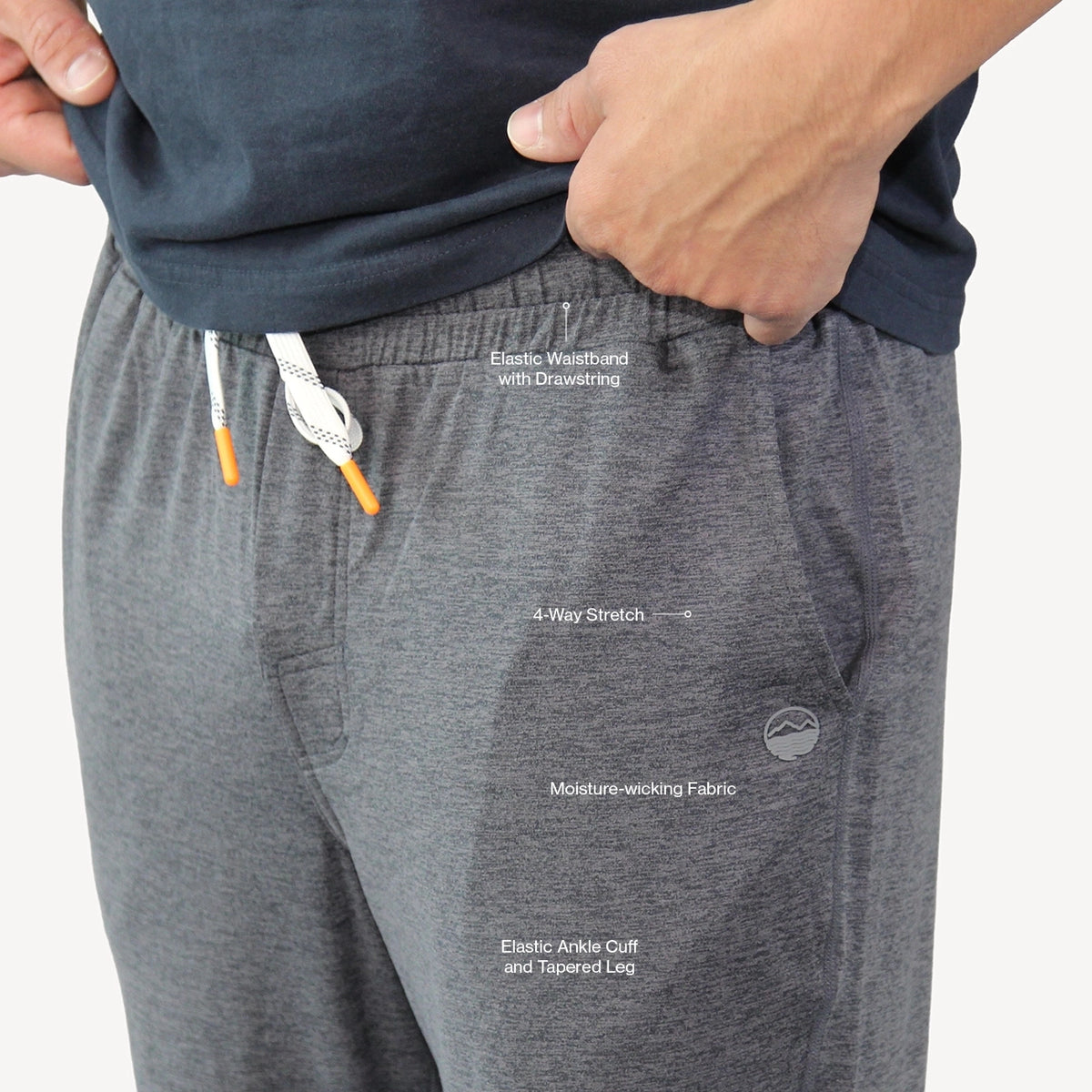 Close-up of a person wearing heather gray drawstring sweatpants with white text callouts highlighting features like elastic waistband with drawstring, 4-way stretch, moisture-wicking fabric, and elastic ankle cuff. The person is also wearing a navy top, and their hands are visible pulling the fabric.