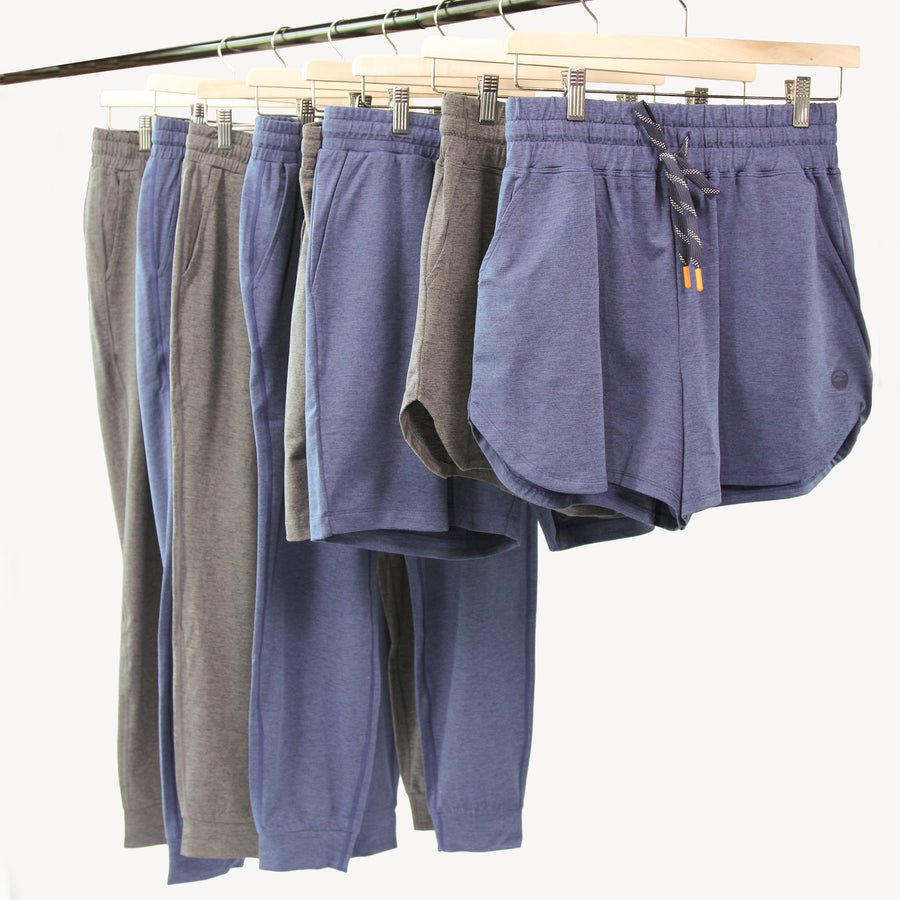 A set of women's and men's lounge shorts in multiple colors (gray, blue, purple, and taupe) hanging on wooden hangers from a metal rack against a plain white background.