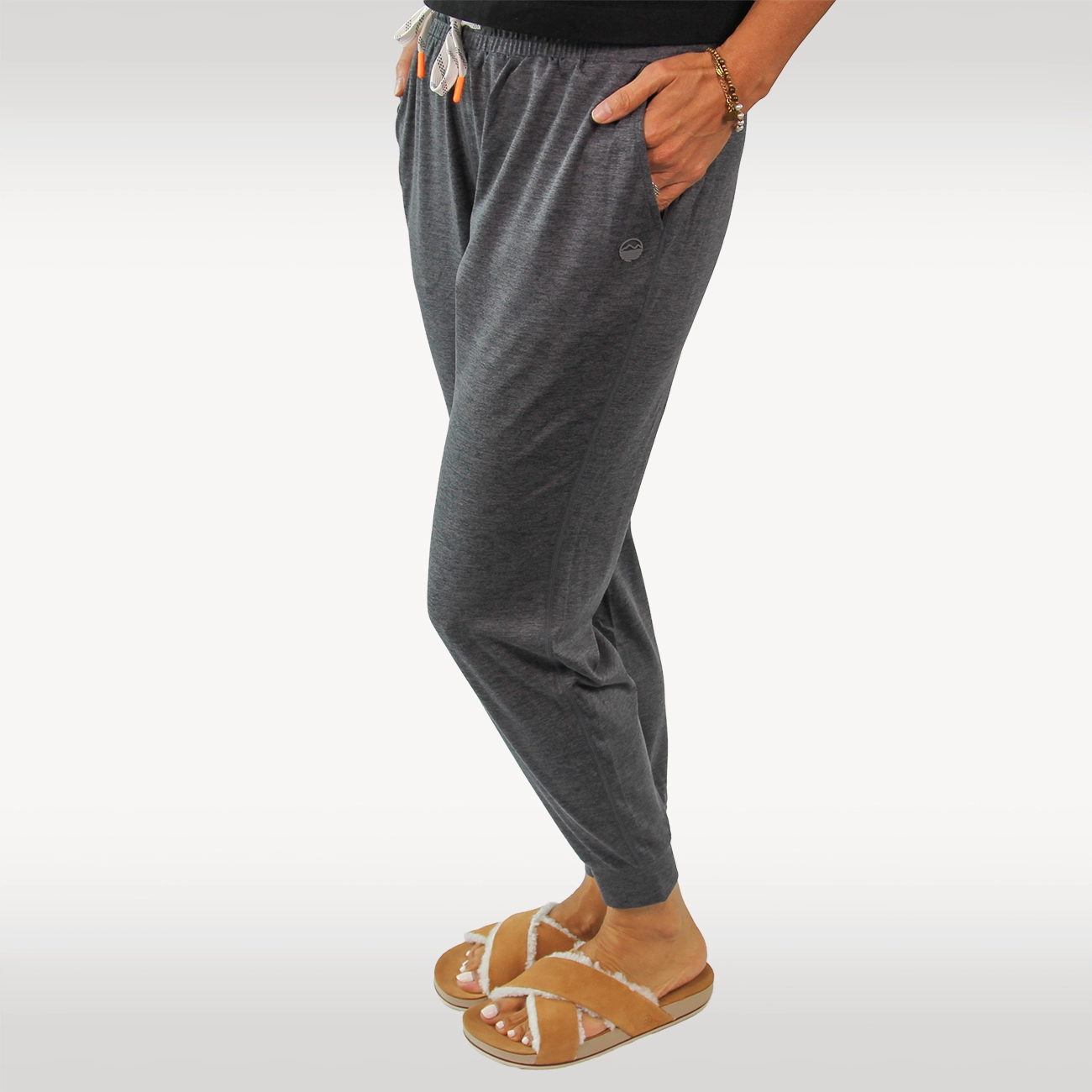 A woman wearing dark heather gray drawstring joggers with side pockets, paired with tan Hari Mari slipers and a black top, shown from the waist down against a light gray studio background.