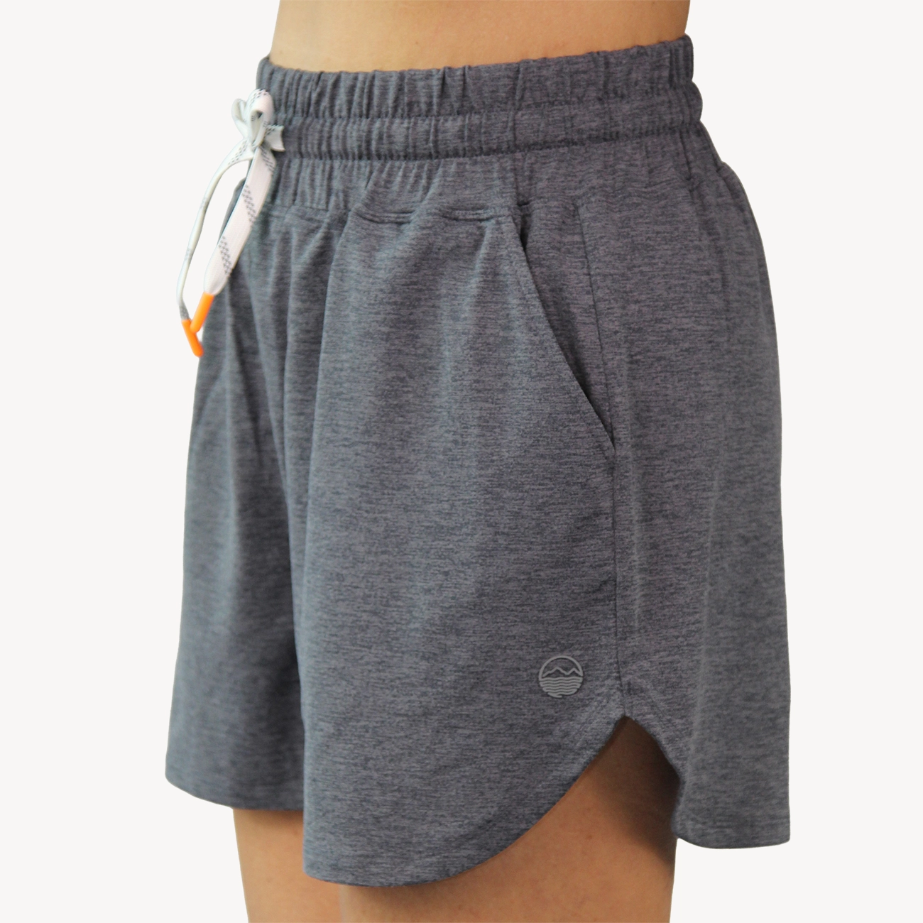 A close-up of a woman wearing heather gray Hari Mari athletic shorts with an elastic waistband, white drawstring, side pockets, and a small circular logo near the lower left leg.
