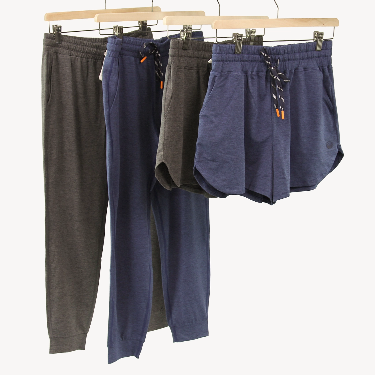 Four pairs of casual lounge bottoms from Hari Mari hanging on wooden hangers against a plain light background: two jogger pants in dark gray and navy, and two shorts in dark gray and navy, each with elastic waistbands and drawstrings.