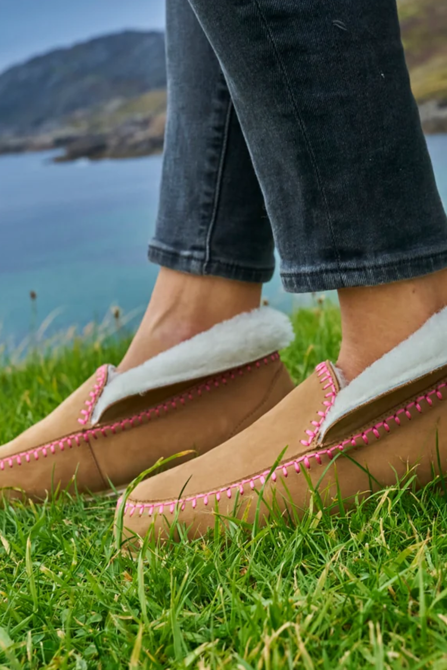A close-up of a person wearing tan suede mule slippers with white shearling lining and pink stitching, standing on green grass with a scenic coastal landscape in the background.