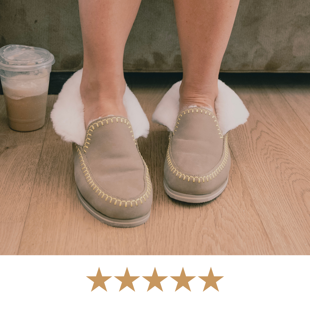 A person wearing taupe slip-on slippers with white  shearling and gold stitching, standing on a light wood floor next to an iced coffee. A five-star graphic overlay is at the bottom.