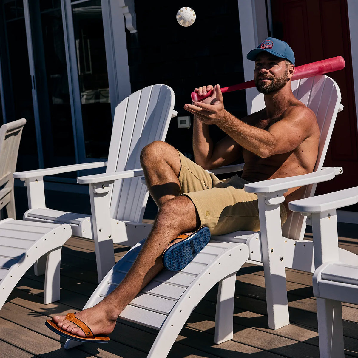 Man sitting on a white Adirondack chair holding a red baseball bat and wearing Nokona x Hari Mari flip flops