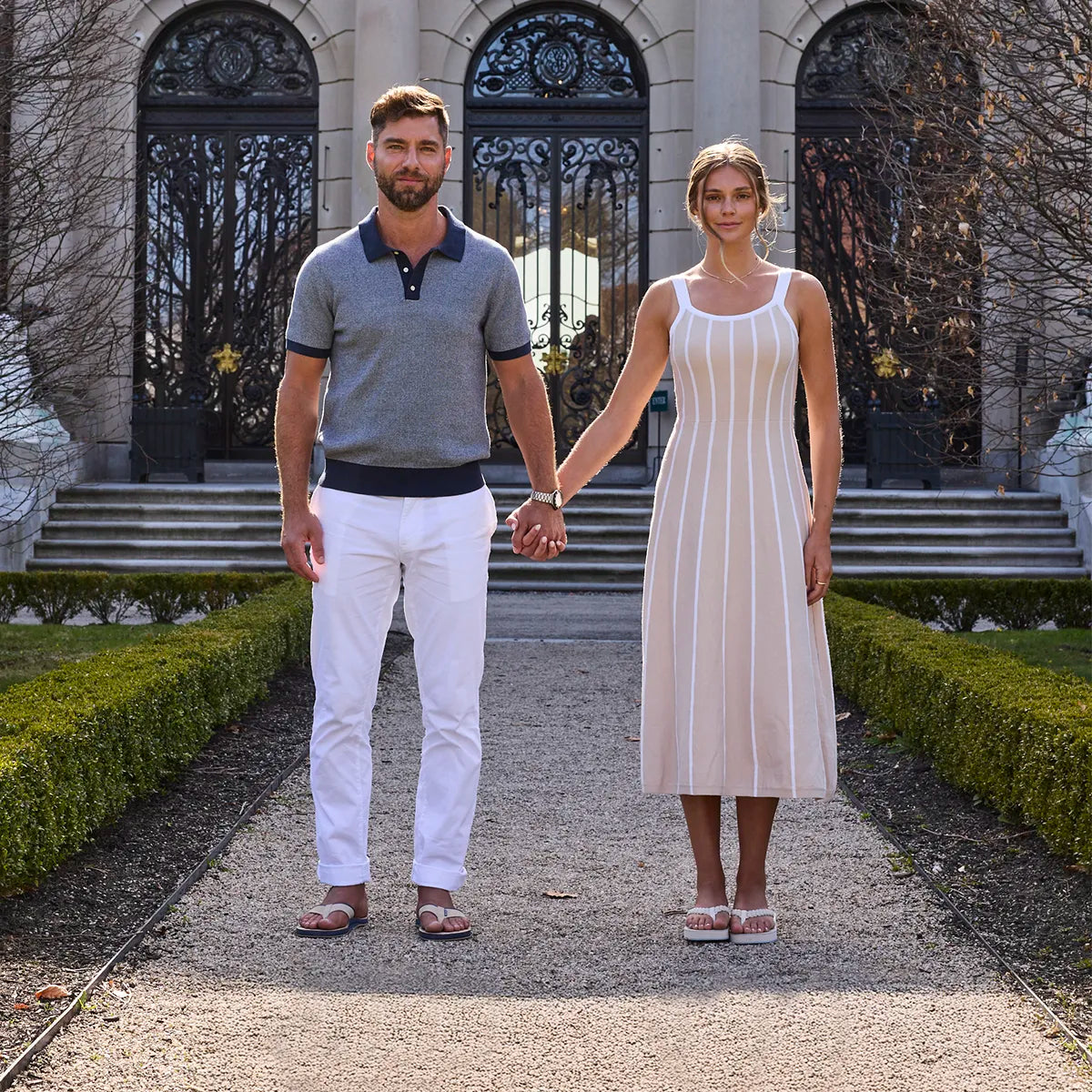 A man and a woman holding hands, standing on a gravel path in front of a grand stone building with large iron gates, both wearing Hari Mari flip flops.